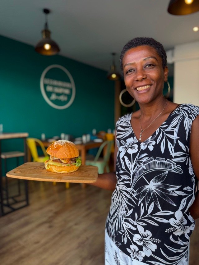 Burger présenté sur une planche dans la salle, devant le mur turquoise et le logo