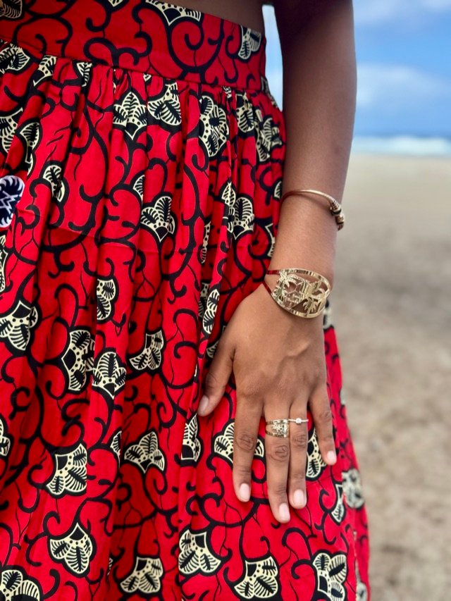 Bracelet jonc et bagues dorés portés sur une jupe en wax rouge, en bord de plage